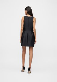 Γυναίκα όρθια, γυρισμένη προς τα πίσω, φοράει μαύρο μίνι φόρεμα χωρίς μανίκια με φερμουάρ στην πλάτη και μαύρα πέδιλα με λουράκι στον αστράγαλο και ψηλό τακούνι.