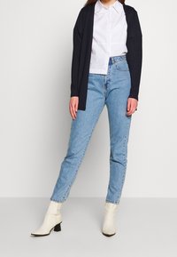Marineblaue Langarm-Cardigan über einem weißen Hemd, kombiniert mit hellblauen, hochgeschnittenen Jeans und weißen Ankle Boots mit Blockabsatz.