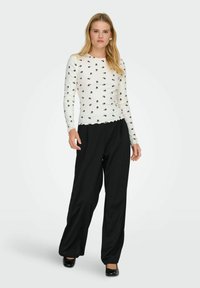 Biały top z długim rękawem, prążkowany, z czarnym wzorem kwiatowym; z falbaniastym wykończeniem. W połączeniu z luźnymi czarnymi spodniami i czarnymi butami.