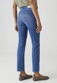 Persoon die blauwe rechttoe-rechtaan jeans draagt, een olijfgroen gebreid mouwloos topje en grijze platte schoenen, staand met ontspannen handen langs het lichaam.