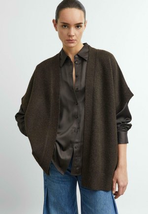 Woman wearing dark brown open-front knit cardigan over a black satin button-up shirt and blue wide-leg jeans against a plain background.