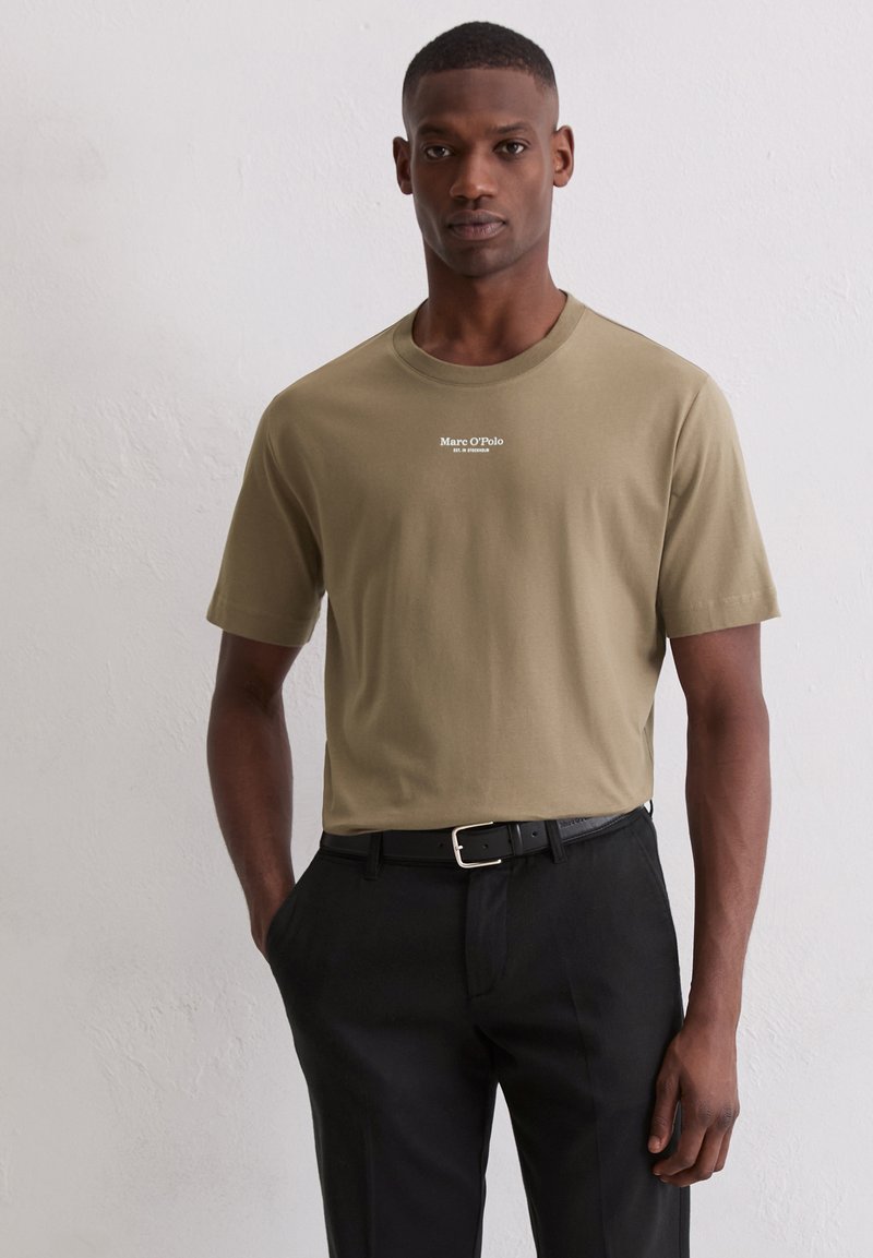 Man in een olijfgroen Marc O'Polo t-shirt dat in zwarte broek is gestopt, met een zwarte riem, staande tegen een eenvoudige witte muur.