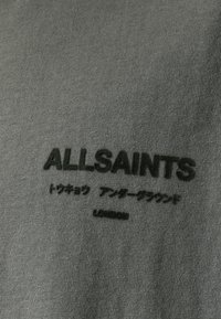 Camiseta de algodón verde oscuro con logo impreso en negro de "ALLSAINTS" y caracteres asiáticos, con un acabado de tela suave y texturizada.
