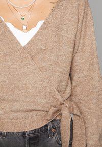 Maglione avvolgente marrone con una texture morbida, con una cintura da annodare in vita. Sotto c'è una maglia bianca con scollatura a onda. Jeans scuri visibili.