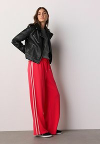 Veste de biker en cuir noir sur un pull gris, associée à un pantalon large rouge vif avec des bandes blanches sur les côtés, porté avec des baskets noires.