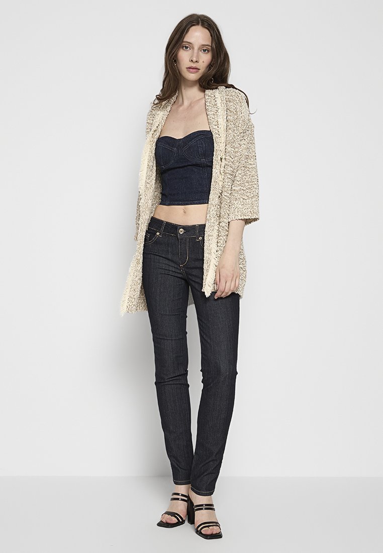 Cardigan beige con texture, top senza maniche in denim scuro e jeans skinny scuri. Sandali neri con cinturini evidenziano l'insieme.