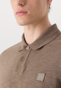 Smeđi polo majica od teksturirane tkanine s klasičnim ovratnikom, zatvaranjem na dva gumba i malom logotipnom oznakom na prsima.