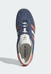 Tenisky Adidas s tmavě modrým semišovým svrškem, bílými tkaničkami a růžovými akcenty. Mají texturovaný bílý jazyk a hnědou gumovou podrážku.