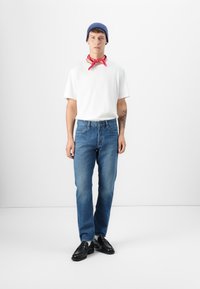 Camiseta blanca, gorro azul, bandana roja, jeans de denim, zapatos negros. El modelo está de pie sobre un fondo blanco, mostrando un diseño de atuendo casual.
