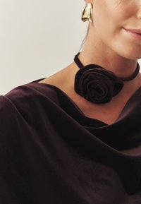 Softe dybe lilla velourstof draperer sig blødt om halsen, med en sort rose detalje. En guld statement-ørering fuldender looket.