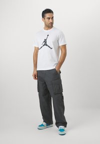 Hvid bomuld T-shirt med en sort grafik af en silhuet i en springpose, parret med mørkegrå cargo-bukser og blå sneakers.