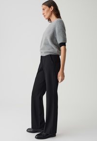 Lichtgrijze fuzzy trui met korte mouwen, gecombineerd met op maat gemaakte zwarte broek. Het model staat zijwaarts, waardoor de pasvorm van de outfit goed zichtbaar is.