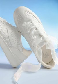 Vita sneakers med rundade tår, ventilationshål och texturerade mönster. Har satinband och en slät gummisula.