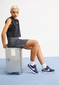 Schwarzes Crop-Top und graue Shorts; das Modell trägt navyfarbene Nike-Sneaker und weiße Socken, sitzt auf einer metallischen Kiste vor einem schlichten Hintergrund.