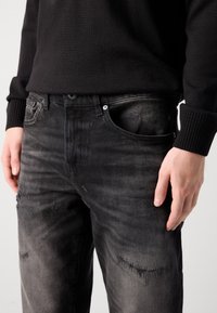 Blugi din denim negru cu un finisaj uzat și distorsionat, având cinci buzunare și închidere cu nasture. Tessut texturat cu variații subtile de culoare.