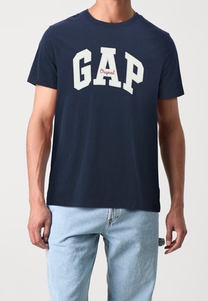 Camiseta de algodón azul marino con gran logo blanco de "GAP" con acentos rojos. Cuello redondo clásico y mangas cortas, ajuste relajado.