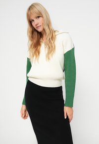 Pull-over en tricot avec un corps crème, des manches côtelées vertes et un col zippé mi-hauteur, associé à une jupe noire ajustée. Tissu lisse et texturé.