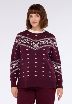 Frau mit schulterlangem braunem Haar, die einen kastanienbraunen Pullover mit weißen Paisley- und Blumenmustern sowie passende Hosen trägt.