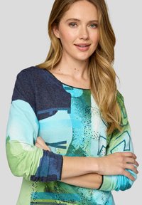 Frau mit langen blonden Haaren, die ein buntes langärmliges Oberteil in Blau-, Grün- und Türkistönen trägt, mit verschränkten Armen und sanftem Lächeln.