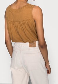 Zandbruine mouwloze top met geplooide achterkant, gecombineerd met hoge taille lichtbeige jeans met zakken en label.