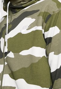 Maskáčová mikina s kapucí v zelené, bílé a tmavě zelené barvě. Má přední zip, kapuci a bílý logo Nike na hrudi. Měkká textura.