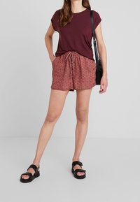 Bordeaux t-shirt med uppvikta ärmar, mönstrade röda shorts med dragsko i midjan och svarta sandaler med öppen design och tjocka remmar.