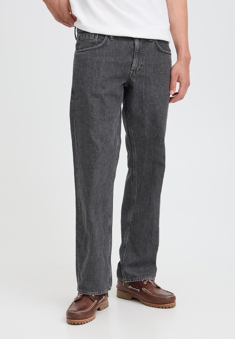 Homme portant un jean gris foncé droit et des chaussures bateau en cuir marron, debout devant un fond clair uni.