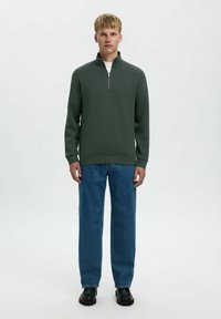Felpa verde con cerniera a quarto di zip e una texture liscia, abbinata a jeans blu larghi e scarpe nere. Design semplice e casual.