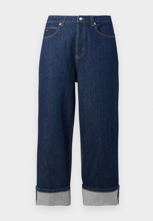 Jeans de perna larga em azul escuro com bainhas dobradas, botão na frente e costura de contraste sobre um fundo branco liso.