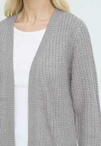 Jasnoszara dziergana kardigan z otwartym przodem, z teksturowanym wzorem i ściągaczami na rękawach, noszony na białej koszulce z okrągłym dekoltem.