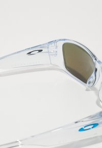 Klare Kunststoff-Sonnenbrille mit dunklen Gläsern, die ein aerodynamisches Design und strukturierte Akzente an den Bügeln aufweist, beschriftet mit "TOURNIQUET."