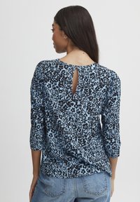 Camicia a stampa floreale blu con maniche 3/4, caratterizzata da un dettaglio a chiave nella parte posteriore. Il tessuto ha una consistenza morbida con motivi floreali neri.