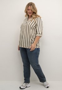 Beige blouse met verticale zwarte strepen, knoopsluiting bij de kraag en opgerolde mouwen, gecombineerd met blauwe denim jeans en lichtgekleurde sneakers.