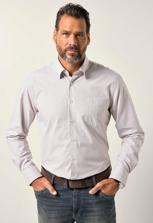 Mann mit Bart, der ein hell gemustertes Hemd, eine blaue Jeans, einen Ledergürtel und eine Uhr trägt, steht mit den Händen in den Taschen vor einem einfarbigen Hintergrund.