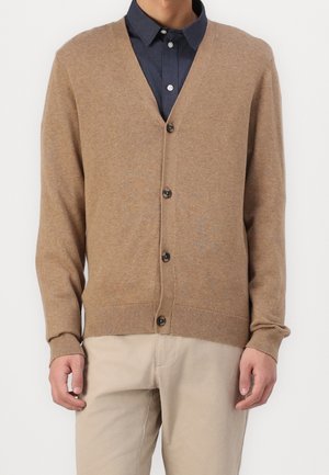 Uomo che indossa un cardigan beige con bottoni sopra una camicia blu navy e pantaloni beige chiaro, mostrato dalle spalle ai fianchi su uno sfondo bianco.