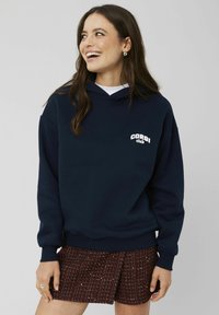Hoodie bleu marine avec une poche frontale et un texte "Corgi Club" en blanc. Tissu doux, coupe décontractée, associé à une jupe bordeaux à motifs.