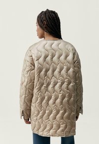 Padded béžový kabát s vlnitým vzorem, volného střihu, dlouhými rukávy a bočními rozparky pro snadný pohyb. Hladká, lesklá látka.