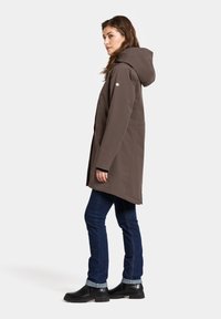 Bruine waterdichte jas met een capuchon, zijzakken en drukknoopsluitingen; gecombineerd met donkere spijkerbroek en zwarte enkellaarsjes.