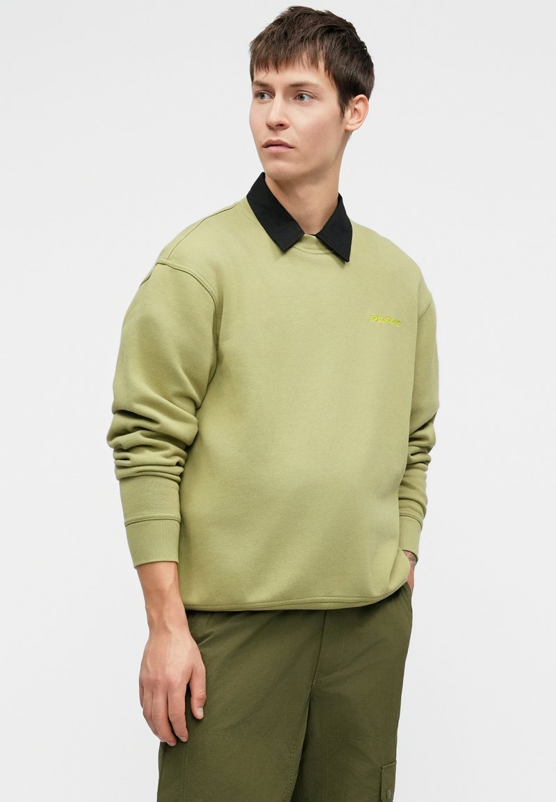 Sudadera verde oliva con un cuello negro, escote redondo y puños acanalados. Presenta un pequeño logo en amarillo brillante en el pecho.