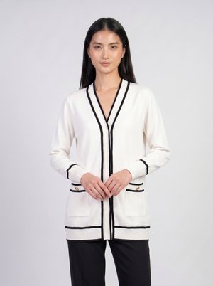 Жена, носеща бял кардиган с черен кант, с дълги ръкави, предни джобове и черни панталони, стои срещу обикновен светъл фон.