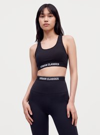 Noor naine, kes kannab musta spordirinnahoidjat ja kõrge vöökohaga retuuse, mõlemad Urban Classics brändi, seisab käed külgedel.