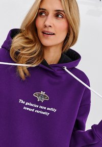 Fialová mikina vyrobená z měkké látky, s černě lemovanou kapucí, bílými šňůrkami a grafickým motivem můry s textem: "Galaxie se jemně otáčejí směrem k zvědavosti."