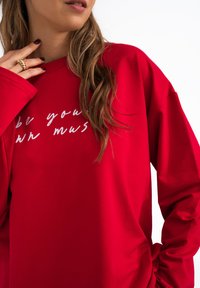 Czerwony, oversize'owy sweatshirt z długim rękawem, z białym, odręcznym napisem na przodzie. Miękka, gładka tekstura i luźny krój.