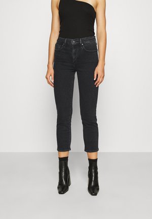 Femme portant un top noir asymétrique, un jean skinny noir taille haute et des bottines noires, debout devant un fond uni.
