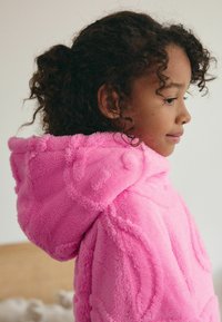 Hoodie en peluche rose avec une surface douce et texturée et une capuche. Affiche un léger brillant et présente des motifs de couture arrondis.