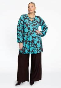 Tunique à manches longues dans un motif floral turquoise sur un fond noir, associée à un pantalon large marron et des talons aiguilles marron. Un collier en chaîne dorée.