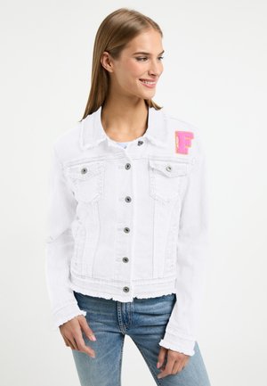 Vrouw draagt een witte spijkerjasje met knoopsluitingen en een roze "F" patch op de linker schouder, gecombineerd met een blauwe jeans.