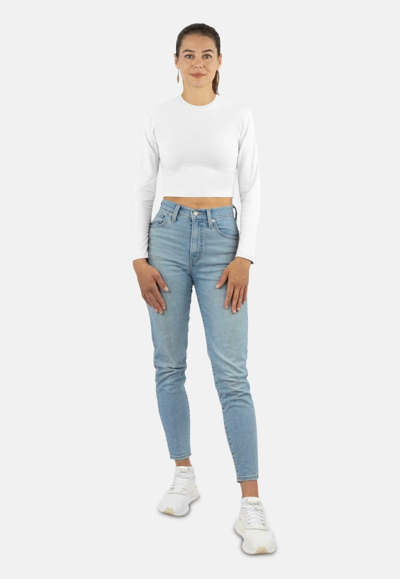 Weißes, langärmliges Crop-Top kombiniert mit hellblauen, hochgeschnittenen Jeans und weißen Sneakers. Einfaches Design, figurbetonte Silhouette, minimale Details.