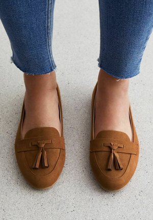 Mocassins en daim marron avec détail de glands ; bout arrondi et semelle plate. Portés avec un jean bleu longueur cheville, mettant en valeur les ourlets effilochés.