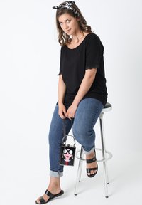 Zwart T-shirt met korte mouwen en kantafwerking, gecombineerd met blauwe jeans en zwarte instapsandalen; houdt een kleine zwarte tas vast met een cartoongezicht.